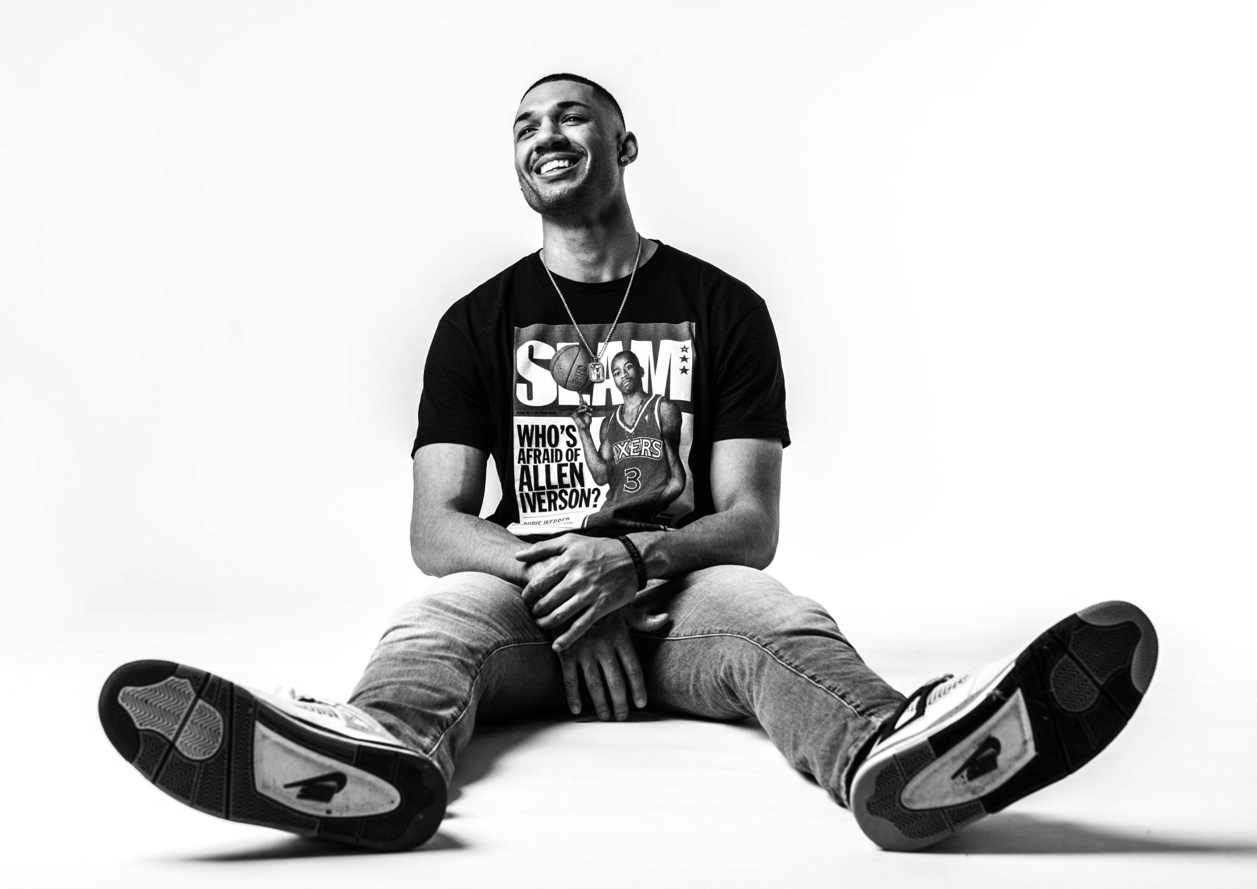 Svartvit porträttbild av en glad ung man i casual stil, poserande på golvet med korslagda ben, iförd en Allen Iverson t-shirt, för en artikel om porträttfotografering.