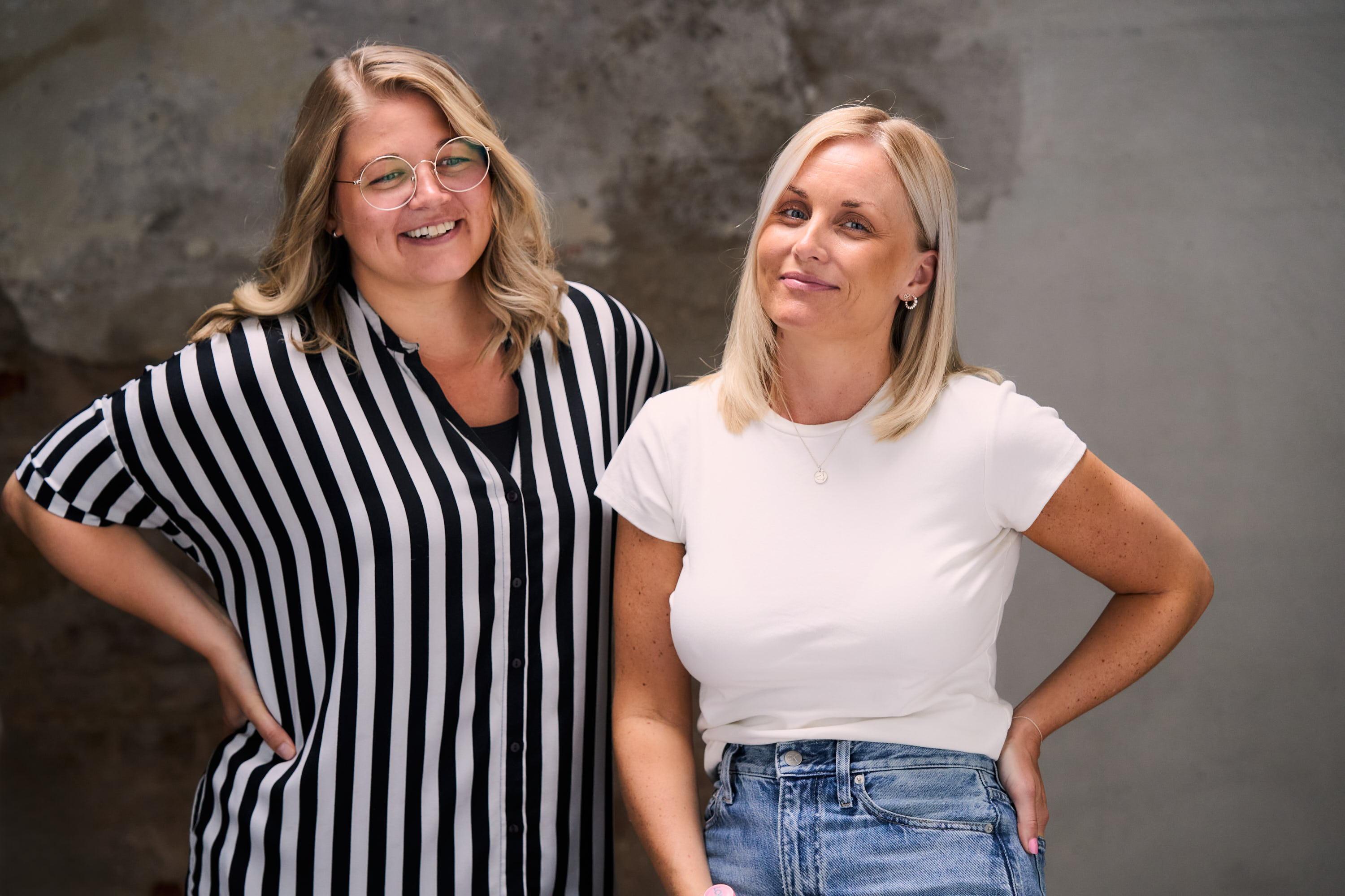 Porträtt av Åsa och Emelie från Yaya Media framför en neutral bakgrund. Åsa, till vänster, bär en svartvit randig skjorta och glasögon, medan Emelie, till höger, har en vit t-shirt och jeans. Båda ler och visar en avslappnad och vänlig attityd.